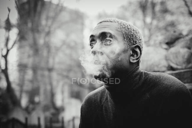 Black and white shot of African American man looking away and exhaling cigarette smoke while resting in city park — Stock Photo