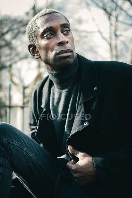 Confident black guy adjusting stylish jacket and looking away while sitting and resting in city park — Stock Photo