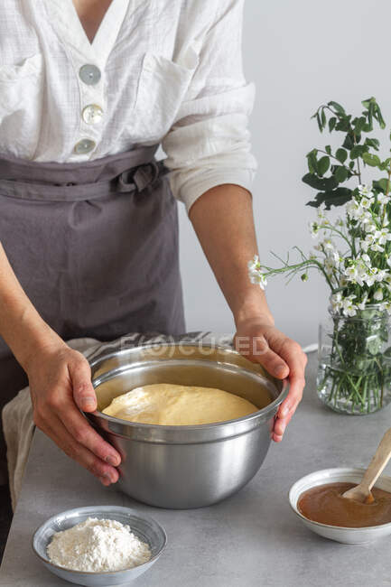 Непізнана жінка в сірому фартусі кладе чашу зі свіжим тістом на стіл біля борошна та яблучного пюре під час приготування тіста — стокове фото