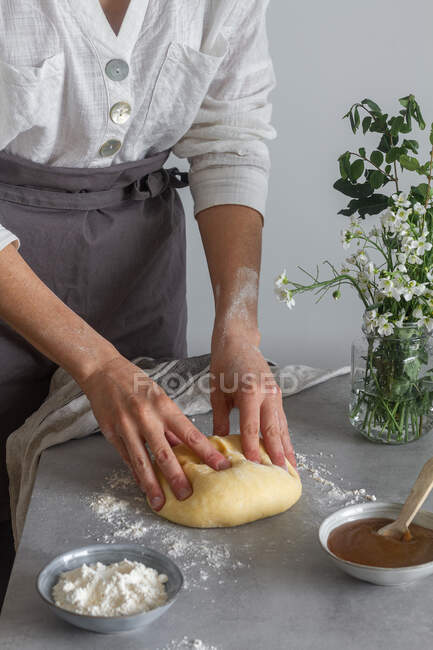 Anonyme Bäckerin in Schürze knetet weichen Teig mit Mehl auf dem Tisch in der Nähe von Apfelmus und Blumenstrauß beim Kochen von Gebäck — Stockfoto