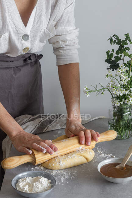 Femme anonyme en tablier roulant pâte à pâtisserie molle sur la table près de la farine et de la compote de pommes fraîches — Photo de stock