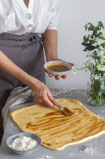 Невпізнавана леді в фартусі, що викладає свіже яблучне пюре з м'якого тіста, готуючи тісто біля борошна і букет квітів — стокове фото