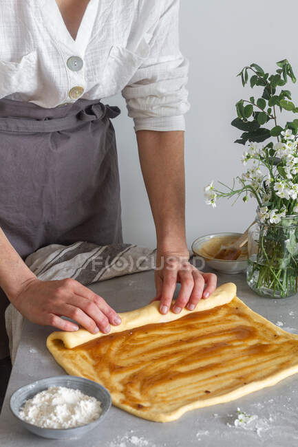 Анонимная пекарь в фартуке делает рулон из мягкого теста с яблочным соусом на столе возле муки и букет цветов — стоковое фото