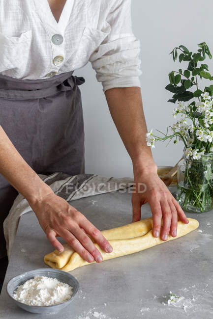 Anonymous woman baker in apron making roll from soft dough on table near flour and bouquet of flowers — Stock Photo