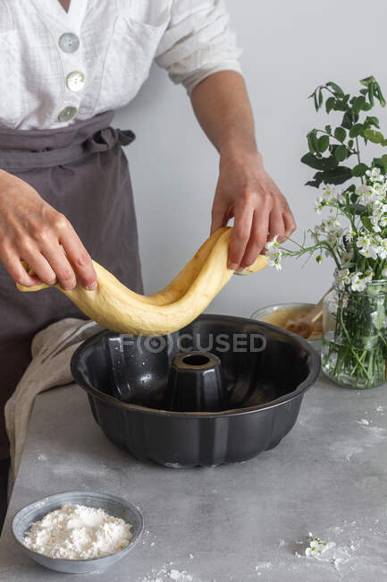 Невизначена жінка в фартусі кладе рулон м'якого тіста на сковороду біля борошна і квітів, готуючи торт Бундт на столі — стокове фото