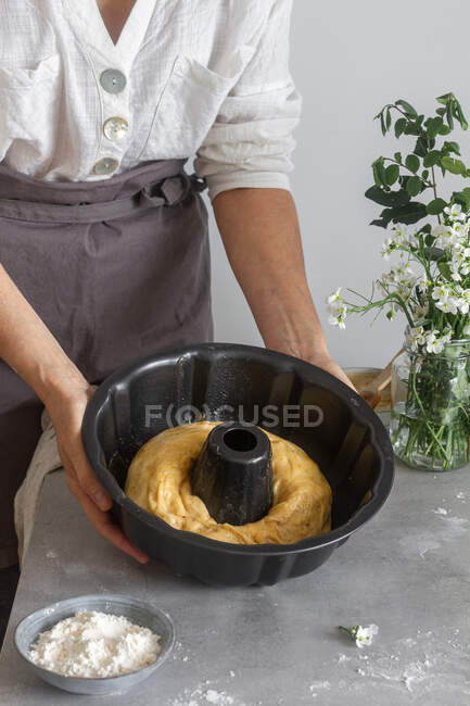 Невизначена жінка в фартусі, що показує рулон м'якого тіста на сковороді біля борошна та квітів, готуючи торт Бундт на столі — стокове фото