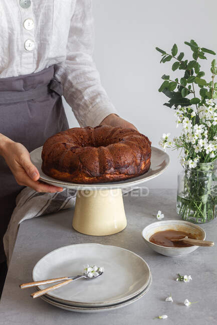 Anonymous female in apron putting plate with fresh Bundt cake on table near flowers and apple sauce — Stock Photo