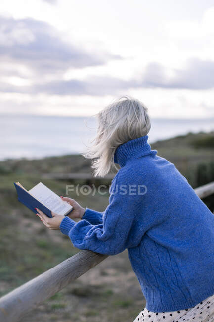 Неузнаваемая молодая девушка в голубом свитере, опирающаяся на деревянный забор и наслаждающаяся любимой поэзией, проводя время на море — стоковое фото