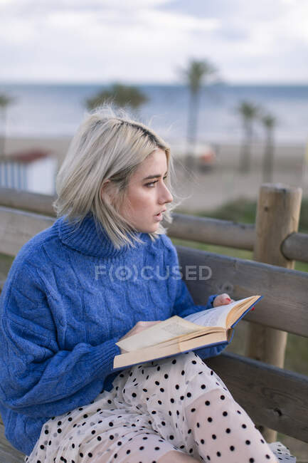 Vista lateral da jovem fêmea loira em suéter azul quente e saia olhando para longe enquanto sentada no banco de madeira no terraço contra a praia turva e livro de leitura — Fotografia de Stock