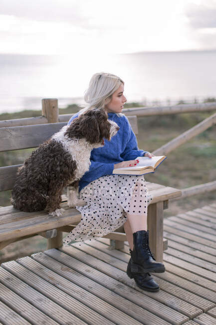 Young female in blue sweater and skirt sitting on wooden bench and petting dog while resting with book at seaside looking away — Stock Photo