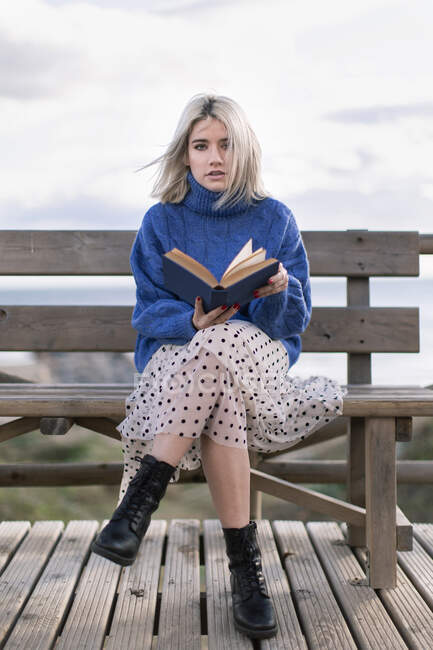 Jeune femme blonde en pull bleu chaud et jupe regardant la caméra assise sur un banc en bois en terrasse contre la plage floue et le livre de lecture — Photo de stock