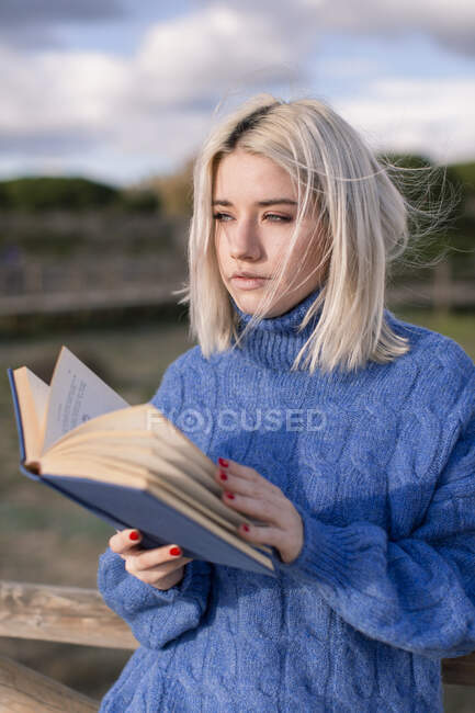 Thoughtful young blonde female in warm blue sweater leaning on wooden fence and reading book while spending spring day in countryside looking away — Stock Photo