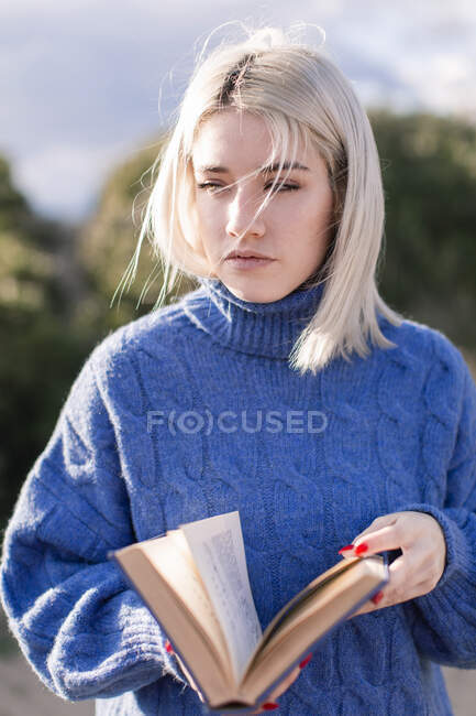 Mujer rubia joven reflexiva en suéter azul cálido apoyándose en una cerca de madera y leyendo un libro mientras pasa el día de primavera en el campo mirando hacia otro lado - foto de stock