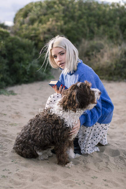 Женщина смотрит на камеру в случайном свитере и юбке с книгой и гладит очаровательную кудрявую собаку, проводя свободное время на песчаном пляже — стоковое фото