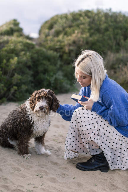Вид сбоку счастливой молодой женщины в обычном свитере и юбке, держащей в руках книгу и гладящей восхитительную кудрявую собаку, проводя свободное время на песчаном пляже — стоковое фото