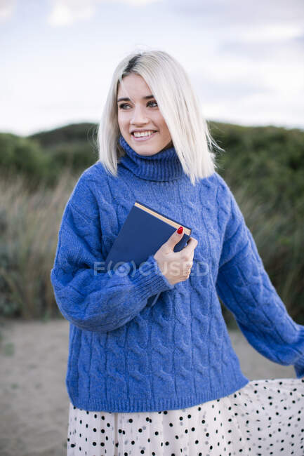 Fröhliche junge Frau mit blonden Haaren im warmen blauen Pullover hält Buch in der Hand und schaut weg, während sie vor verschwommenem natürlichen sandigen Hintergrund steht — Stockfoto
