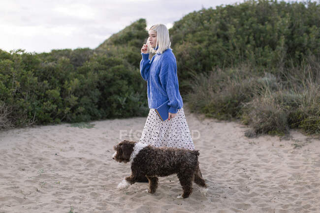 Vista laterale della giovane bionda femmina in maglione casual e gonna con libro in mano passeggiando con cane sulla spiaggia sabbiosa vicino a piante verdi — Foto stock