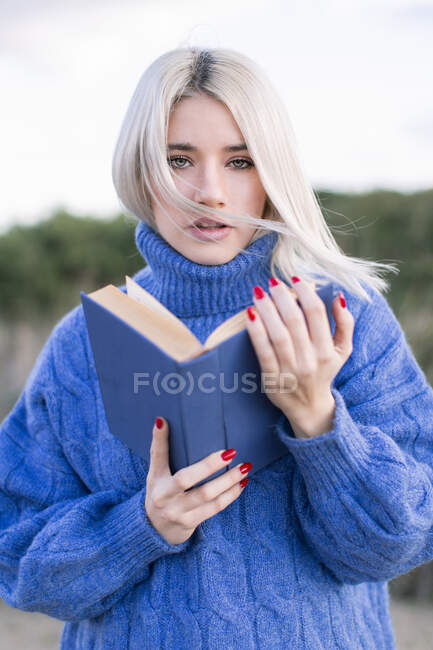 Продумана молода жінка з світлим волоссям у теплому синьому светрі тримає відкриту книгу і дивиться на камеру, стоячи на розмитому природному піщаному фоні — стокове фото