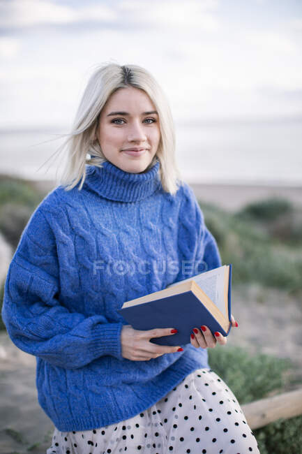 Cheerful young blonde female in warm blue sweater sitting on wooden fence and reading book while spending spring day in countryside looking at camera — Stock Photo