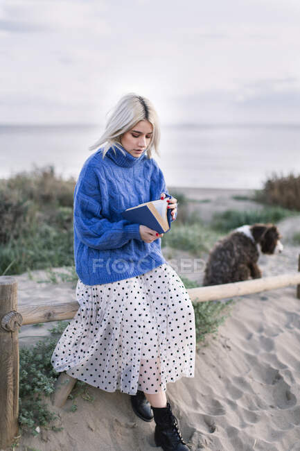 Thoughtful young blonde female in warm blue sweater sitting on wooden fence and reading book while spending spring day in countryside with loyal dog — Stock Photo