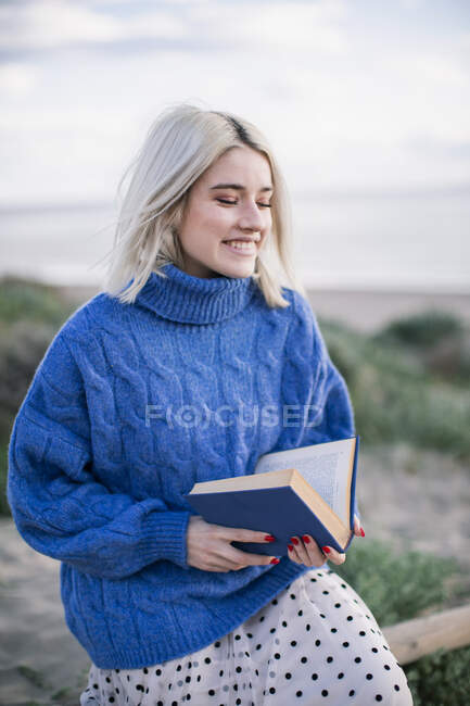Joven rubia alegre con suéter azul cálido sentada en una valla de madera y leyendo un libro mientras pasa el día de primavera en el campo con los ojos cerrados - foto de stock