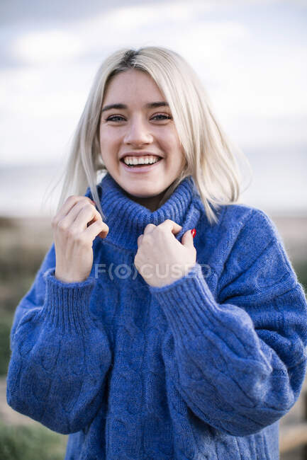 Joyful cheerful young blonde female in blue knitted sweater looking at camera and laughing while standing against blurred background of sea coast — Stock Photo