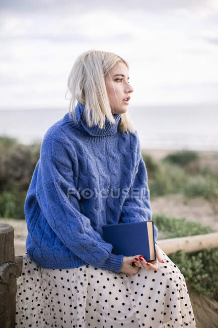 Mujer rubia joven reflexiva en suéter azul cálido sentado en valla de madera sosteniendo libro mientras pasa el día de primavera en el campo mirando hacia otro lado - foto de stock