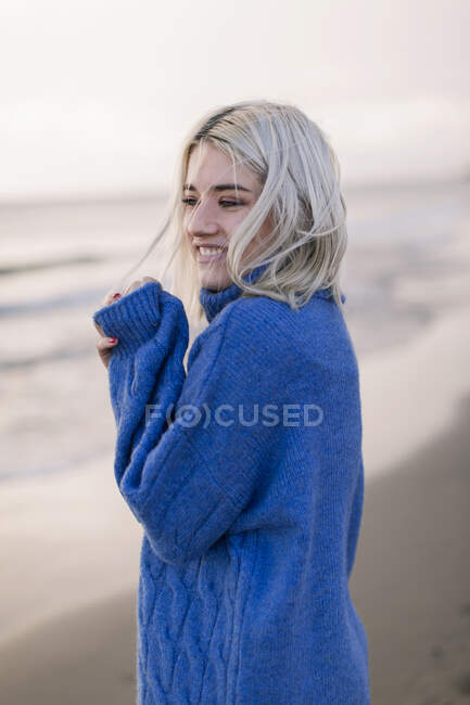 Vista lateral de alegre joven rubia alegre en suéter de punto azul mirando hacia otro lado y riendo mientras está de pie sobre el fondo borroso de la costa del mar - foto de stock