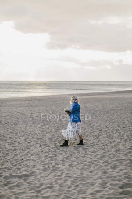 Side view of lonely young female in stylish outfit walking with open book in hand on empty sandy seashore in cloudy day — Stock Photo