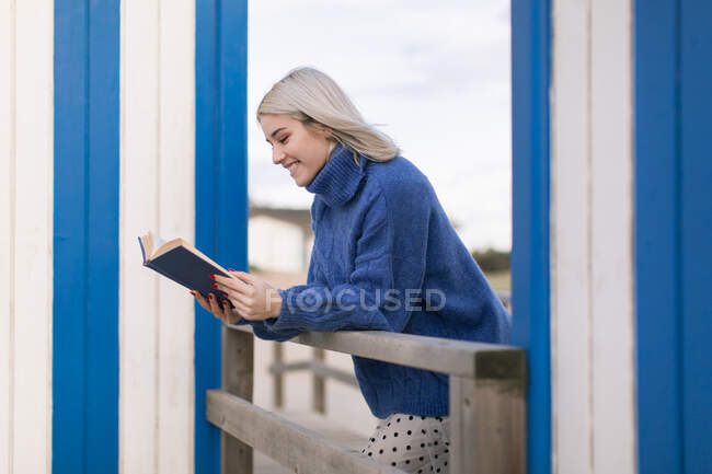 Fröhliche junge Frau in warmem Pullover und Rock, angelehnt an einen Holzzaun mit aufgeschlagenem Buch an weiß-blau gestreifter Wand — Stockfoto