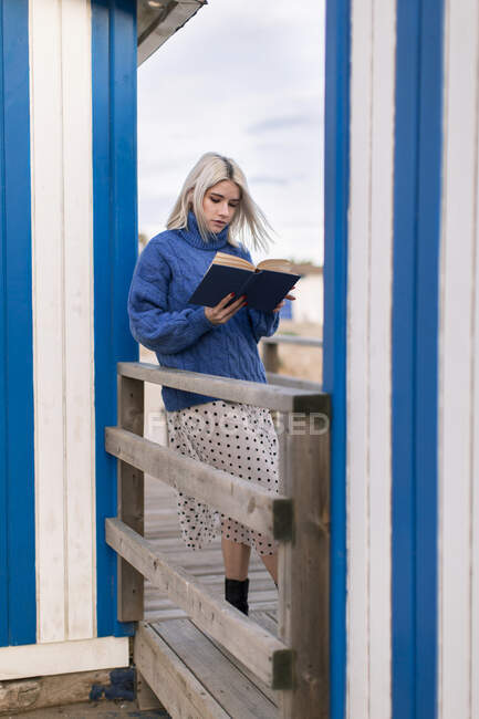 Jeune femme réfléchie en pull chaud et jupe appuyée sur une clôture en bois avec lecture de livre ouvert contre un mur rayé blanc et bleu — Photo de stock