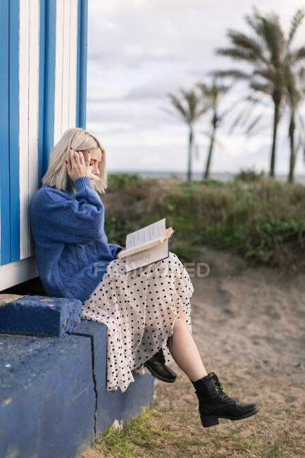Vista laterale della giovane donna pensierosa in maglione caldo e gonna seduta con lettura a libro aperto contro la parete a strisce bianche e blu guardando altrove — Foto stock