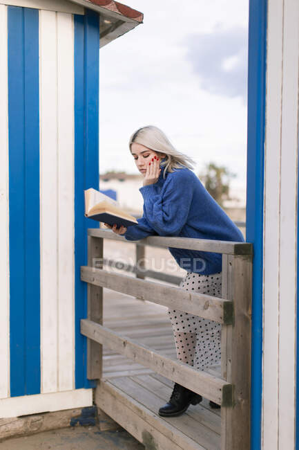 Mujer joven pensativa en suéter cálido y falda apoyada en valla de madera con lectura de libro abierto contra pared de rayas blancas y azules - foto de stock