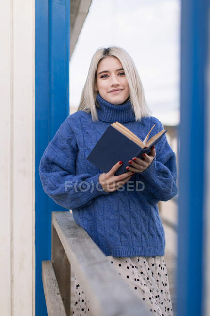 Thoughtful young female in warm sweater and skirt smiling and looking at camera while leaning on wooden fence with open book against white and blue striped wall — Stock Photo