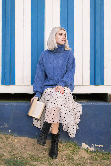 Von unten sitzt eine nachdenkliche junge Frau in warmem Pullover und Rock mit aufgeschlagenem Buch vor weiß-blau gestreifter Wand und schaut weg — Stockfoto