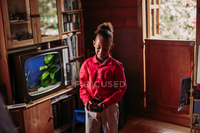 Положительный черный подросток в повседневной одежде с джойстик контроллер в руках играет в комнате с деревянным интерьером и старомодным телевизором — стоковое фото