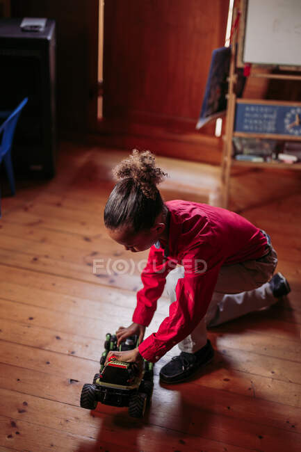 From above side view of ethnic child in casual clothes sitting on wooden floor and playing with toy car at home — Stock Photo