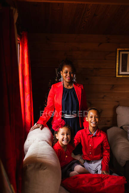 Jovem alegre preto fêmea com bonito rindo irmãos em roupas vermelhas olhando para a câmera enquanto passar o tempo juntos em casa de campo acolhedor — Fotografia de Stock
