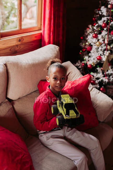 Ragazza nera soddisfatta che tiene in mano il giocattolo e guarda la fotocamera mentre si siede sul divano vicino alla finestra in un accogliente soggiorno con decorazione di Natale — Foto stock