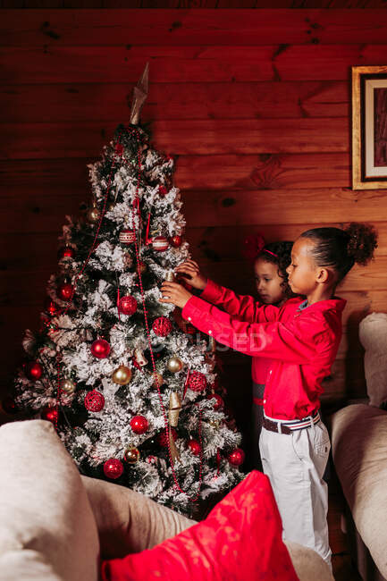 Seitenansicht der niedlichen ethnischen Geschwister in lässiger Kleidung schmücken traditionellen Weihnachtsbaum in gemütlichen Holzhaus — Stockfoto