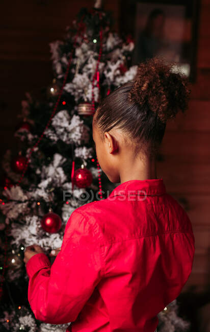 Rückansicht des fröhlichen kleinen schwarzen Mädchens im roten Hemd, das in die Kamera blickt und lächelt, während es neben dem geschmückten Weihnachtsbaum zu Hause steht — Stockfoto