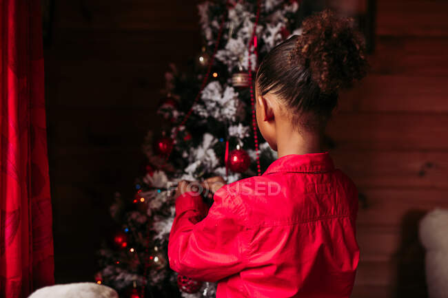 Вид ззаду весела маленька чорна дівчинка в червоній сорочці дивиться на камеру і посміхається, стоячи біля прикрашеної ялинки вдома — стокове фото