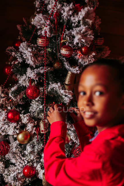 Вид збоку весела маленька чорна дівчинка в червоній сорочці дивиться на камеру і посміхається, стоячи біля прикрашеної ялинки вдома — стокове фото