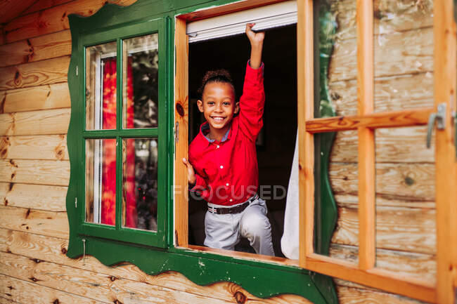 Веселый черный ребенок в красной рубашке и белых брюках смотрит в камеру через открытое окно деревенского деревянного дома — стоковое фото