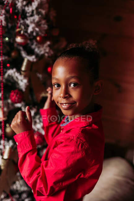 Вид сбоку жизнерадостной маленькой черной девочки в красной рубашке, смотрящей в камеру и улыбающейся, стоя у украшенной елки дома — стоковое фото