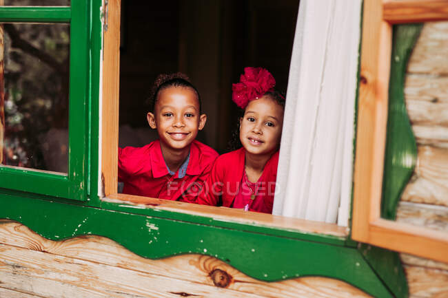 Cute black little siblings smiling and looking at camera through open window of wooden cabin — Stock Photo