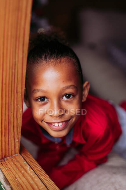 Сверху симпатичная маленькая кудрявая этническая девочка с красным бантиком в красном платье, смотрящая в камеру и улыбаясь на размытом фоне — стоковое фото