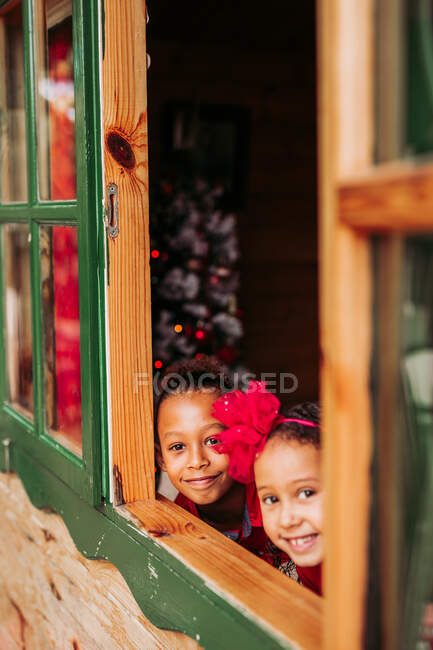 Niedliche schwarze kleine Geschwister lächeln und blicken durch das offene Fenster der Holzhütte in die Kamera — Stockfoto