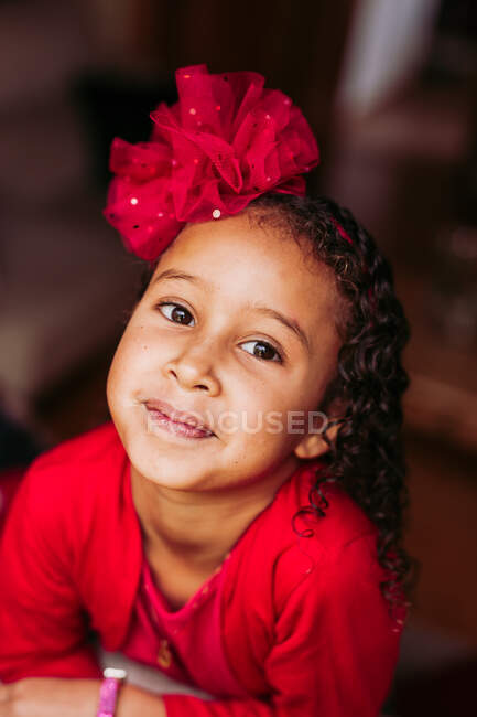Dall'alto della graziosa bambina dai capelli ricci etnici con fiocco rosso che indossa un vestito rosso guardando la fotocamera e sorridendo sullo sfondo sfocato — Foto stock