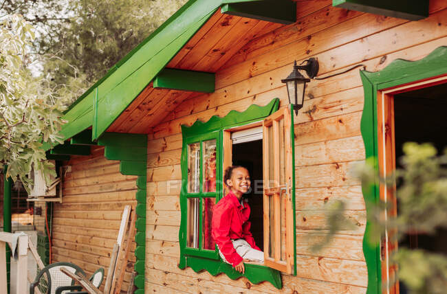 Fröhliches schwarzes Kind in rotem Hemd und weißer Hose blickt durch das offene Fenster eines ländlichen Holzhauses — Stockfoto
